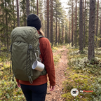 Kvinna går på en stig i tallskog med en silverfärgad aluminiummugg hängande i en karbinhake på ryggsäckens sida