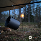 Mörkgrå aluminiummugg som hänger i handtaget på en S-krok under en presenning vid skymning med en lykta i bakgrunden