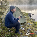 Man sitter på en liten campingpall framför ett grönt tält vid en sjö i morgondimma och håller en mörkgrå aluminiummugg med ånga