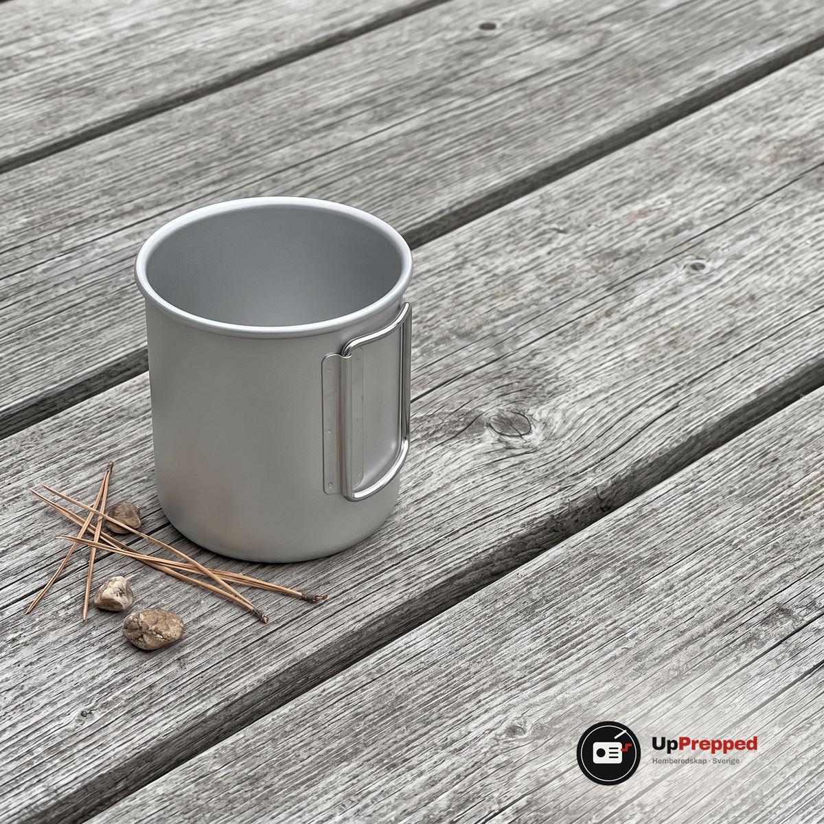 Silverfärgad aluminiummugg med handtaget invikt mot koppen på ett grått träbord med tallbarr och två småstenar