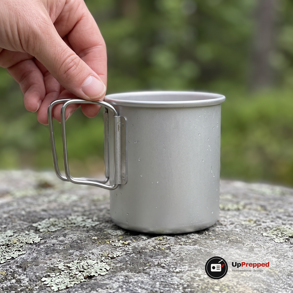 Hand som greppar det vikbara handtaget på en silverfärgad aluminiummugg på en lavbevuxen sten, vattendroppar på ytan