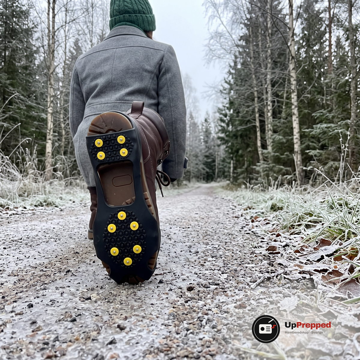 En kvinna går bort längs en frostig grusstig i skogen, den bakre kängans sula är lyft och visar broddar med gula dubbhättor