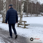 En man i marinblå dunjacka och mössa går på en isig gångväg vid en frusen sjö med broddar under skorna