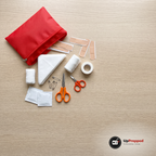 Röd första hjälpen-väska med öppen dragkedja på ett ekbord med plåster, förband, triangelförband, två små saxar, tejp och säkerhetsnålar bredvid