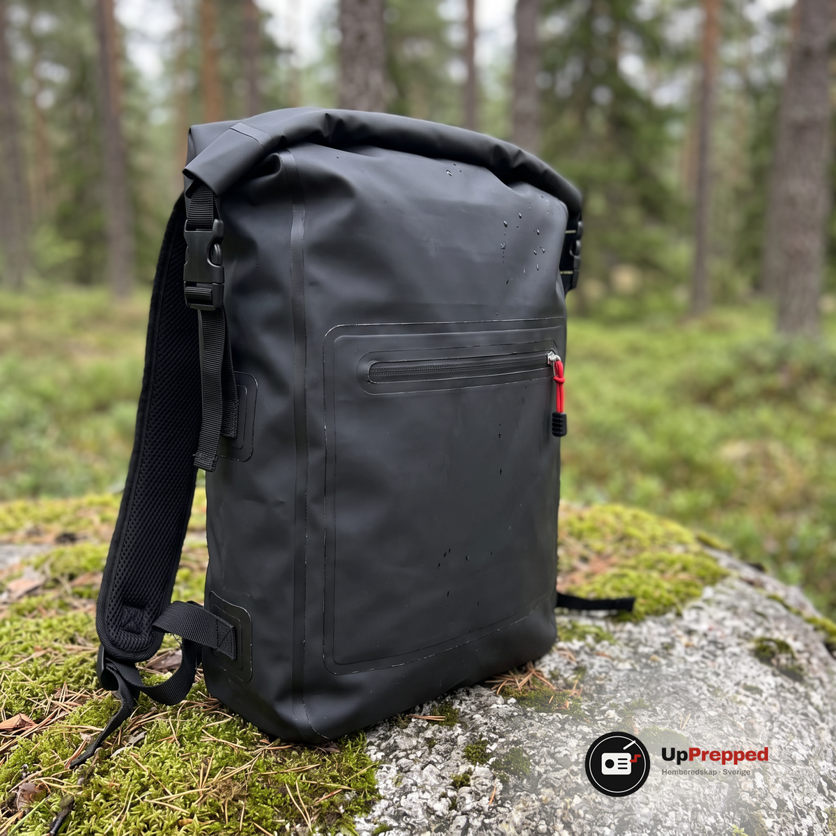 Närbild på den svarta ryggsäckens rulltopp, sidospänne och framficka med röd dragkedjesnodd på en mossig sten i skogen