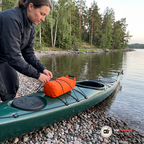 Kvinna justerar elastiska snören över en orange drybag på fördäcket av en grön havskajak på en strand