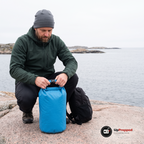 Man i grön softshelljacka rullar ihop toppen på en blå drybag på en granithäll vid havet, en liten ryggsäck står bredvid