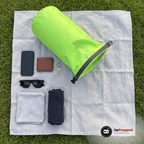Grön drybag öppen på en filt på gräset med mobil, plånbok, nyckel, solglasögon, handduk och t-shirt