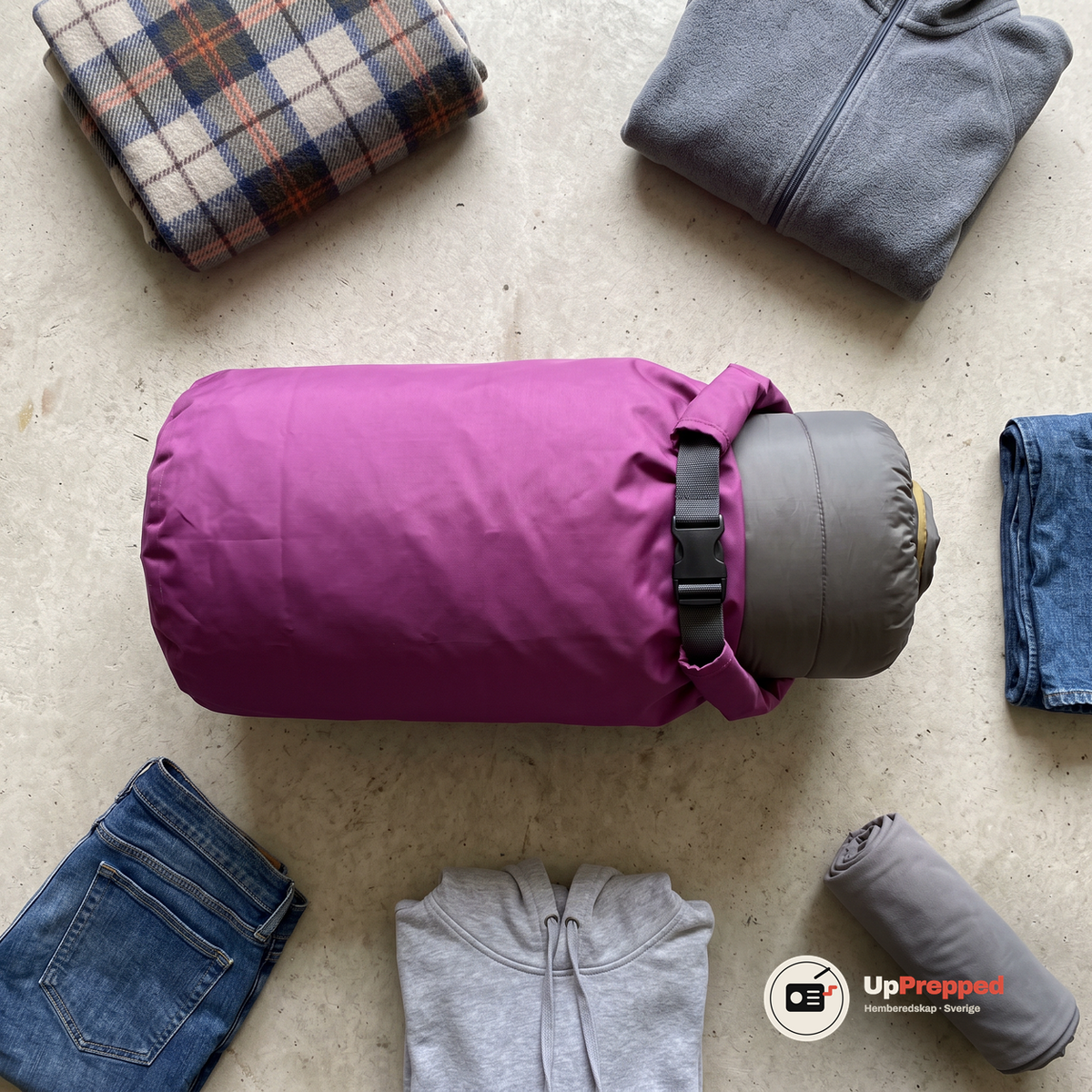 Lila drybag på sidan på ett betonggolv med sovsäck, filt, fleece, jeans, luvtröja och handduk runt omkring