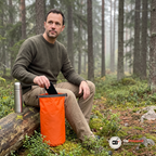 Man på en fallen tall i en svensk skog tar upp en mobil och en nyckel ur en orange drybag, en termos står bredvid
