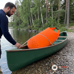 Man i det grunda vattnet kontrollerar repet över en orange drybag som ligger i en grön träkanot på en sjöstrand