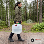Kvinna i olivgrön softshelljacka bär en vattenfylld ihopfällbar vattendunk i det gröna handtaget längs en grusväg vid en campingplats i skogen