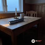 Tänd ficklampa ligger på ett träbord i en stuga i skymningen bredvid en mugg och ett ljus