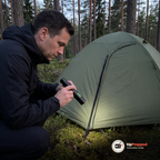 Man hukar vid ett grönt tält i skogen och lyser med ficklampan mot tältdörren och mossan