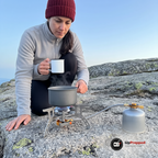 Kvinna i grå fleecetröja och röd mössa kokar vatten i en liten kastrull på gasbrännaren på en bergsklippa, med gaspatronen bredvid