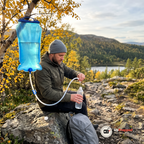 Man i grå mössa och grön jacka sitter på en sten vid en fjällstig och håller en flaska under slangen på den hängda vattensäcken
