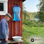 Äldre man i blå arbetsskjorta och keps står vid en röd skjulvägg där vattensäcken hänger på en krok och vatten rinner från slangen ner i ett vitt emaljfat