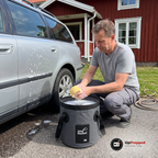 Man på huk vid framhjulet på en silverfärgad kombi sköljer en svamp i hinken fylld med såpvatten