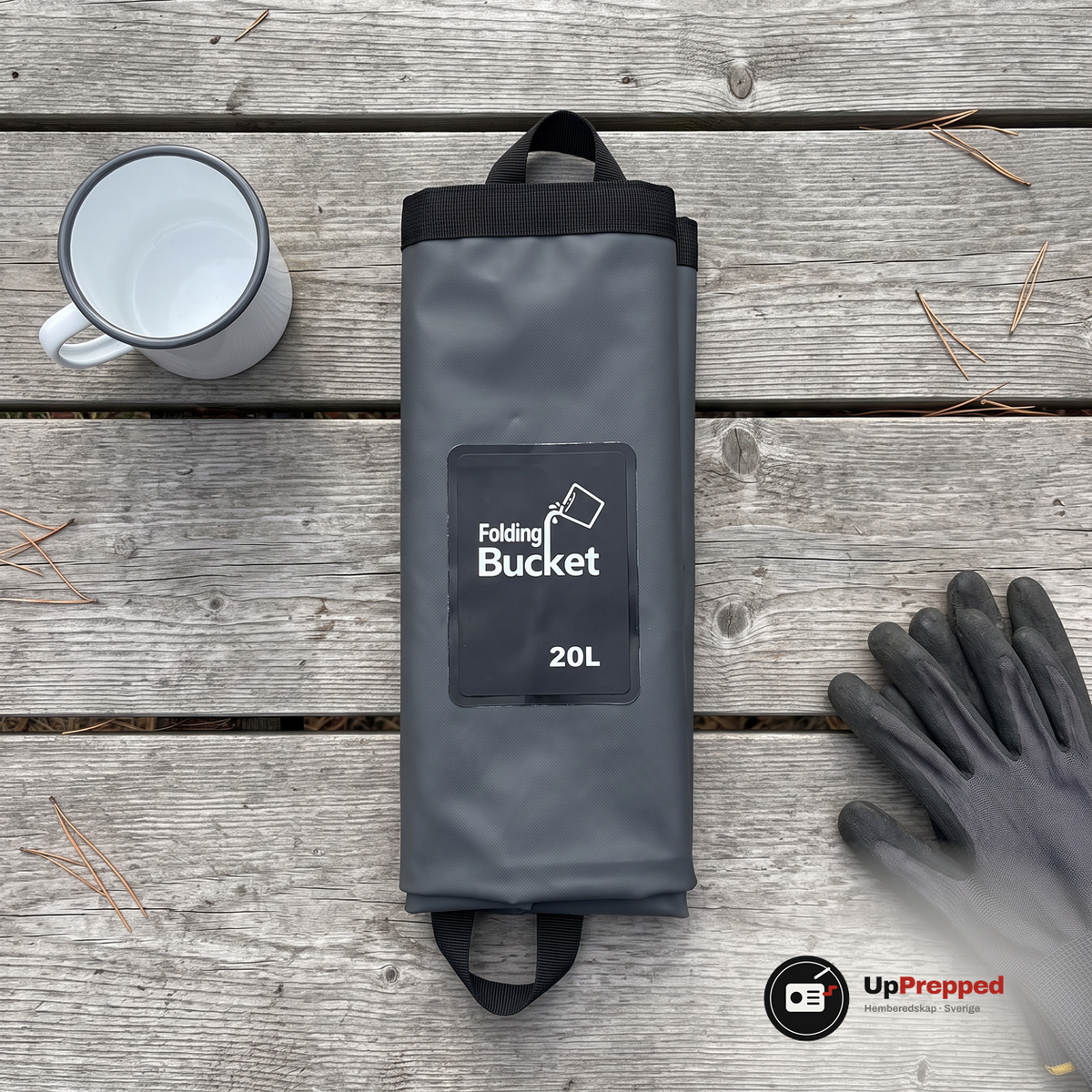 Ihopvikt hink med etiketten Folding Bucket 20L på ett grått träbord bredvid en emaljmugg och ett par handskar