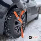 Hand i grå yllehandske håller i den orange remmen på en hjulbrodd vid framhjulet på en parkerad bil