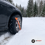 Framhjulet på en mörk bil på en snöig skogsväg med en hjulbrodd och orange rem fäst på däcket