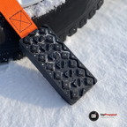 Närbild på ett svart block med upphöjda klackar och orange rem som ligger i snön intill ett däck