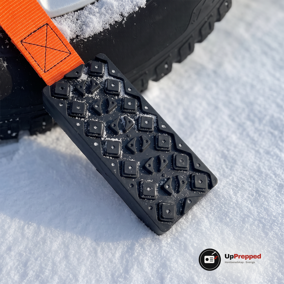 Närbild på ett svart block med upphöjda klackar och orange rem som ligger i snön intill ett däck