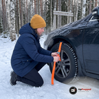 Kvinna knäböjer vid framhjulet på en bil vid en snöig parkering i skogen och spänner den orange remmen på en hjulbrodd