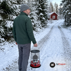 Man i grön jacka bär den släckta, kalla fotogenkaminen i handtaget på en snöig grusväg mot en röd stuga