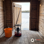 Släckt fotogenkamin bredvid en orange hink på golvet i en trästuga vid en isfiskeplats, dörren öppen mot en frusen sjö och ett metspö lutat mot väggen