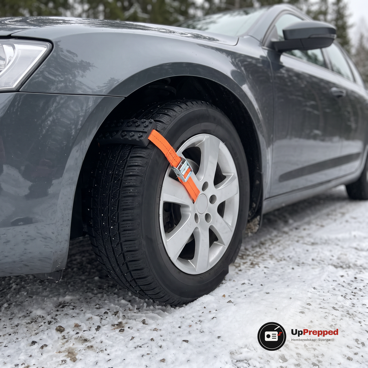 Hjulbrodd med orange rem och spänne fastspänd över däcket på en bil på en snöig grusväg
