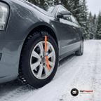 Hjulbrodd med orange rem och spänne fastspänd över däcket på framhjulet på en bil på en snöig grusväg