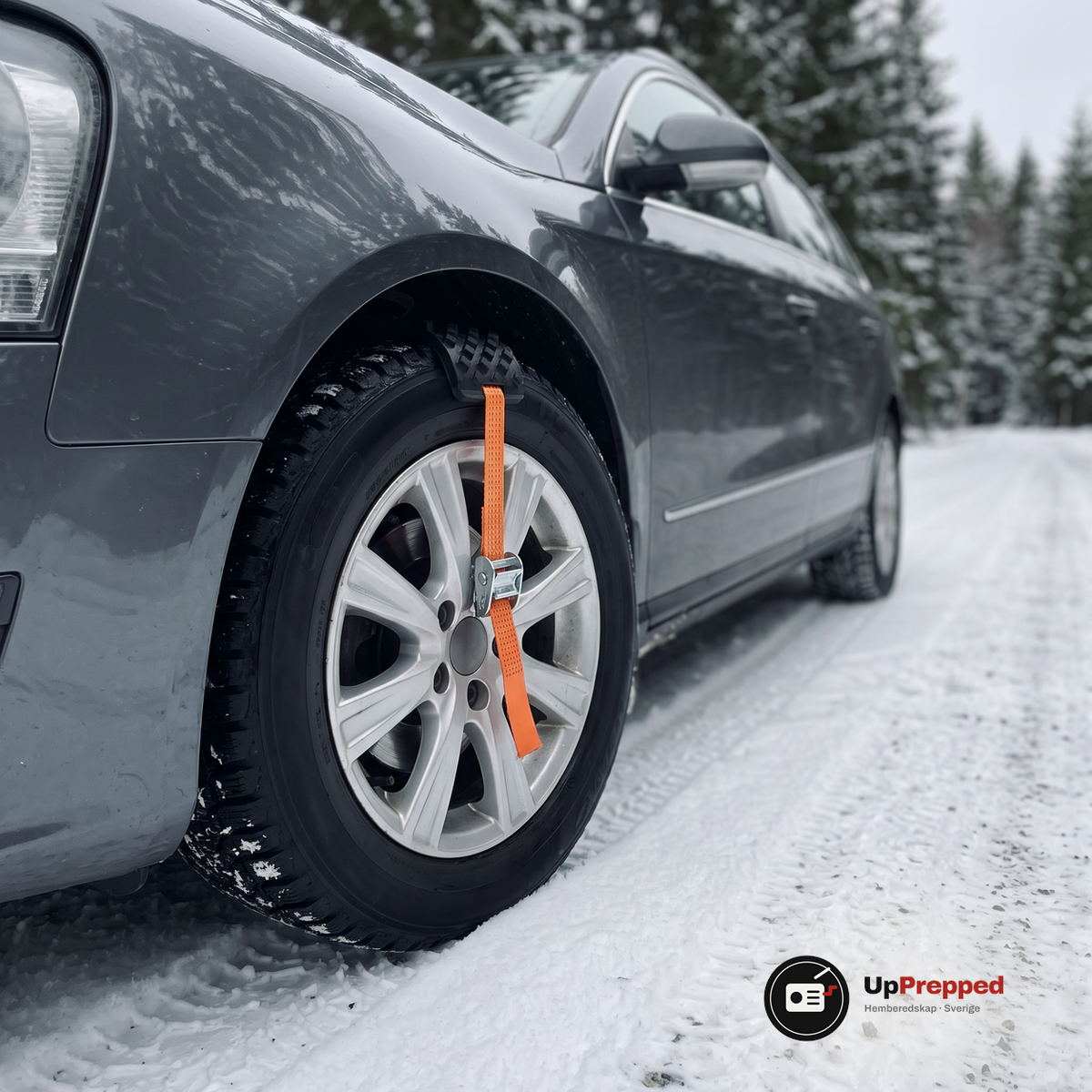 Hjulbrodd med orange rem och spänne fastspänd över däcket på framhjulet på en bil på en snöig grusväg