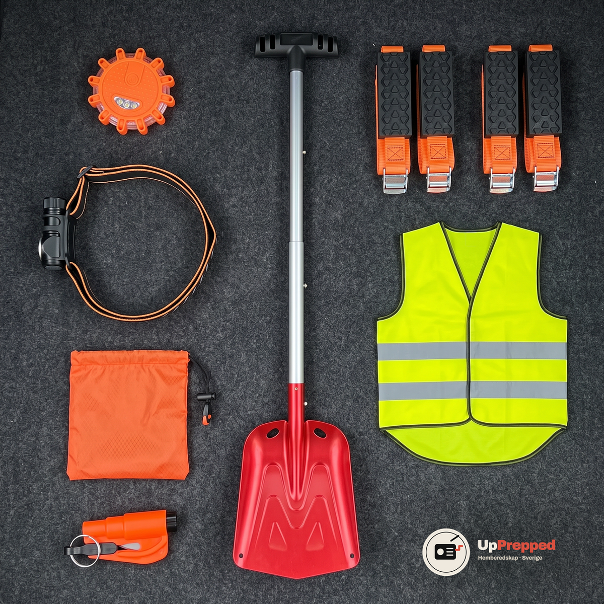 Nödpaketets alla delar uppifrån på ett mörkgrått bagagegolv: varningsljus, pannlampa, nödsovsäck, fönsterkross, snöskyffel, reflexväst och fyra hjulbroddar