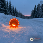 Orange LED varningsljus med magnet, tänt med orange sken, på snön vid kanten av en vinterväg i skymningen