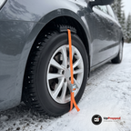 Hjulbrodd med orange rem och spänne fastspänd över däcket på en bil på en snöig grusväg