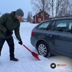 Man i grön jacka skyfflar bort snö vid bakhjulet på en bil med en röd teleskopisk snöskyffel