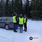 Tre personer i gula reflexvästar står bredvid en parkerad bil på en snöig landsväg i skymningen