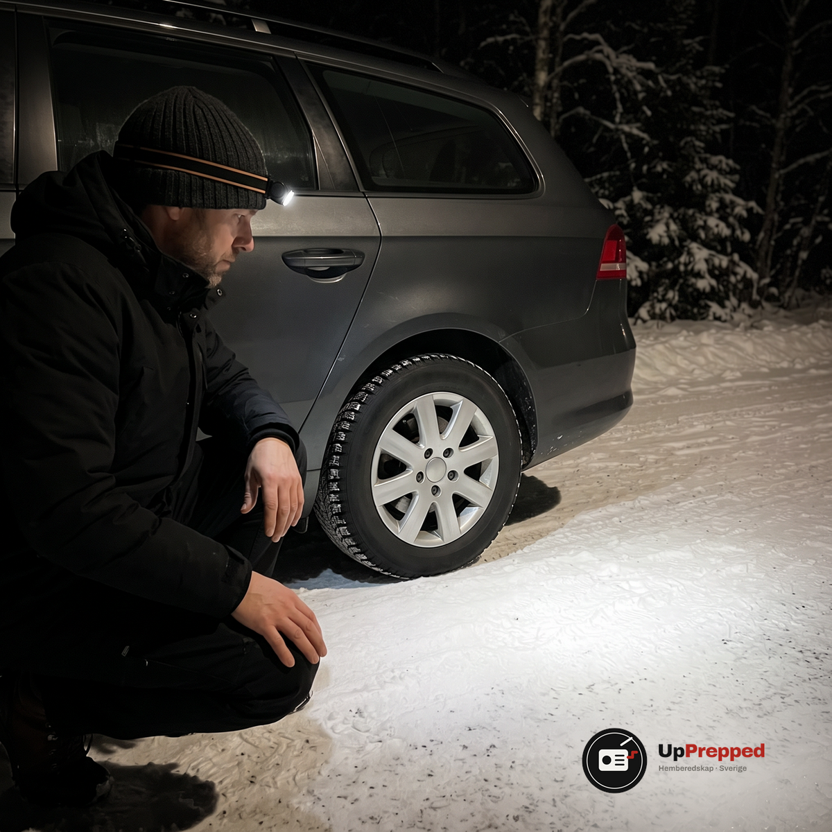 Man med tänd pannlampa på mössan hukar vid bakhjulet på en bil en natt på en snöig landsväg