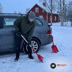 Man i grön jacka skyfflar snö vid bakhjulet på en bil med en röd teleskopisk snöskyffel, en andra snöskyffel lutar mot stötfångaren