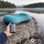 Campingkudden uppblåst på en sten vid en sjö och en hand som håller den lilla grå packpåsen