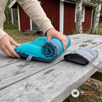 Två händer rullar ihop den tömda campingkudden på ett bord utanför en röd stuga med packpåsen bredvid