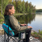 En kvinna i en campingstol vid en sjö med campingkudden som ryggstöd bakom ländryggen