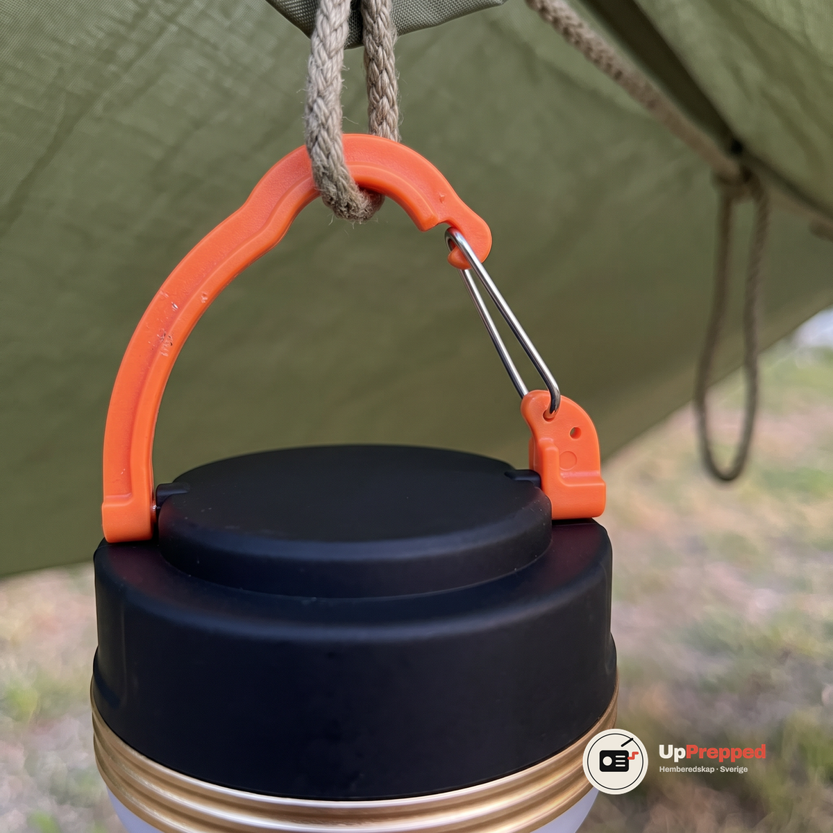 Närbild på lampans orange handtag med silverfärgad klämma fäst i ett rep under ett tarp