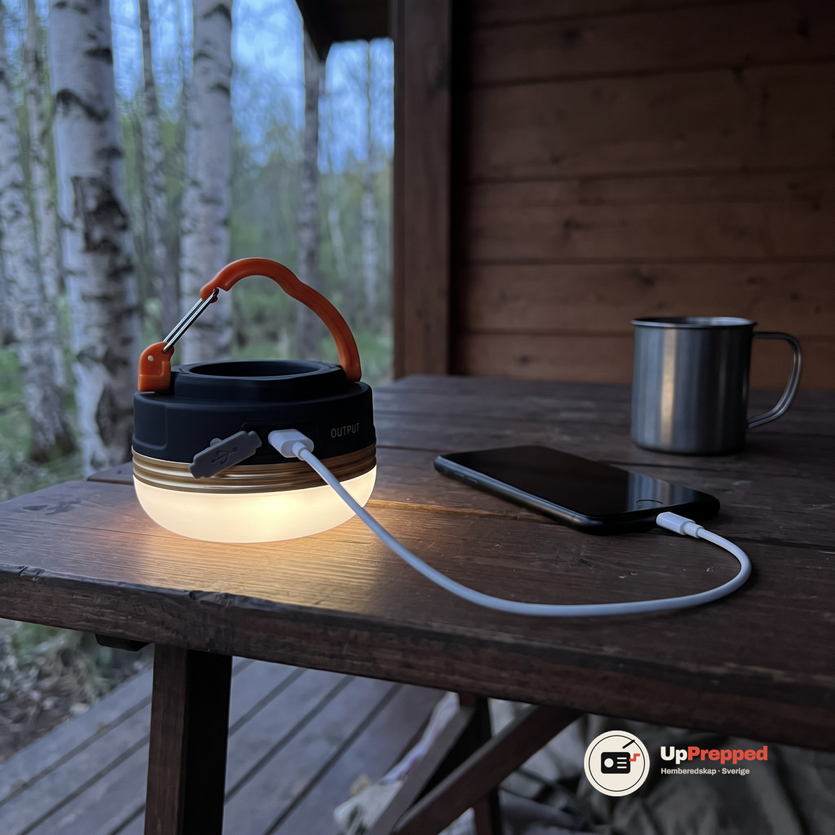 LED campinglampa på ett träbord på en veranda med en vit USB-kabel kopplad till en mobil