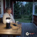 Kvinna i krämfärgad kofta sitter vid ett bord på en veranda i skymningen med lyktan tänd på bordet och en kabel från lyktan till en mobil som laddas