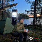 Kvinna i olivgrön fleecejacka med en vit emaljmugg sitter vid ett tält vid en sjö i skymningen medan lyktan hänger tänd i en gren