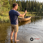 Man står barfota i vattnet vid en skogssjö och håller upp mobilfickan med mobil för att ta en bild av sjön