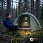 Man sitter på en campingstol vid ett grönt tält i skymningen, dosan hänger tänd under tälttaket