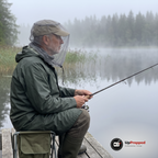 Äldre man i kakifärgad keps med ett grått myggnät över kepsen sitter på en fällstol på en brygga och fiskar en dimmig morgon